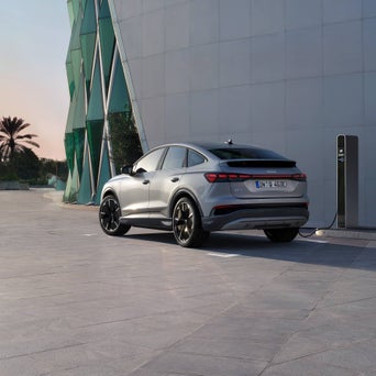 Rückansicht eines hellgrauen Audi Q4 Sportback e-tron, der an einer Schnellladestation im Freien aufgeladen wird, mit einem modernen Gebäude im Hintergrund.