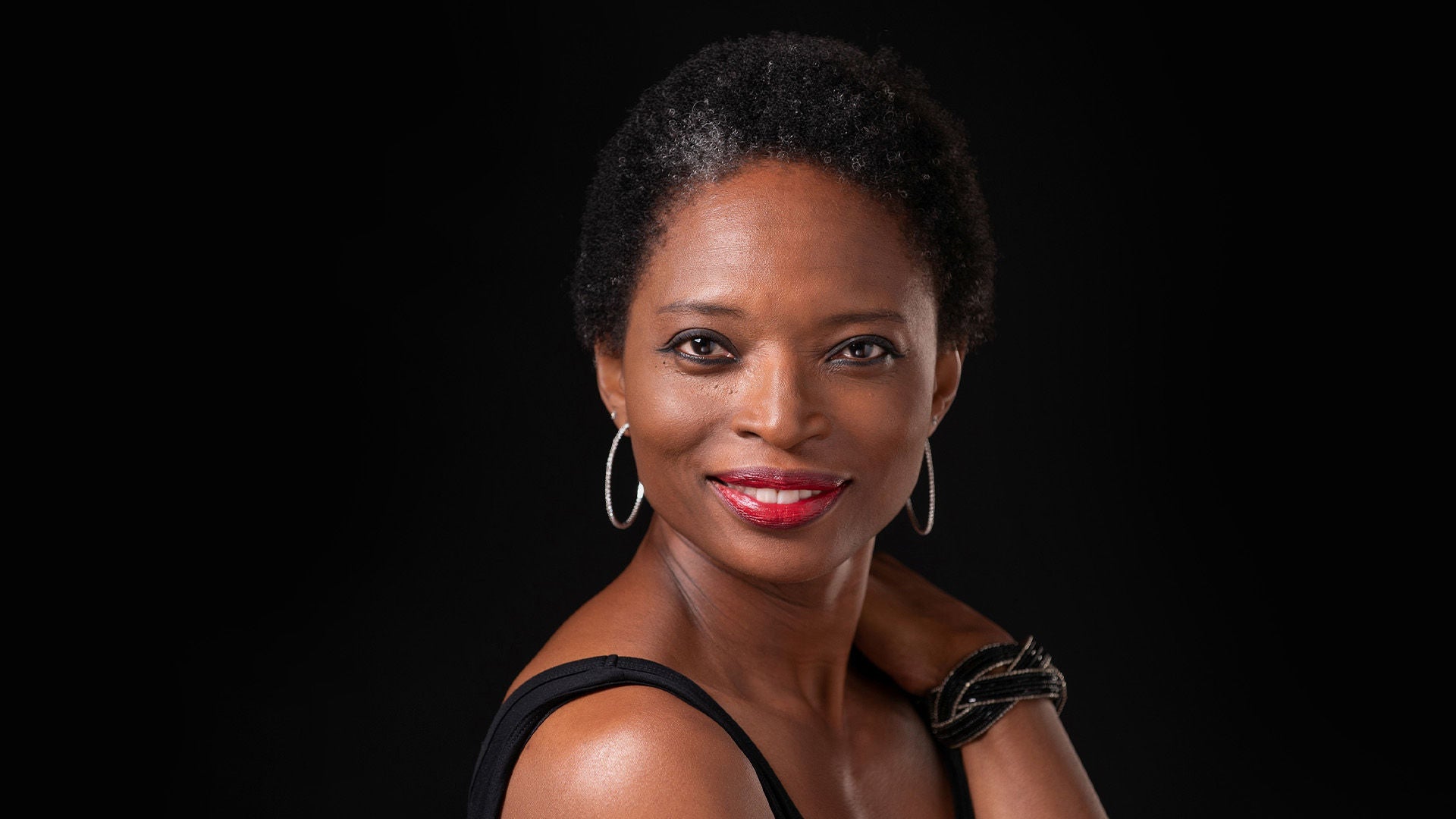 Porträt einer lächelnden Frau mit kurzem Haar, roten Lippen und Creolen; schwarzes Oberteil vor dunklem Hintergrund.