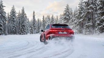Rear view of a red Audi RS 5 Avant on a snow-covered road; swirling snow, tire tracks, and a forest.