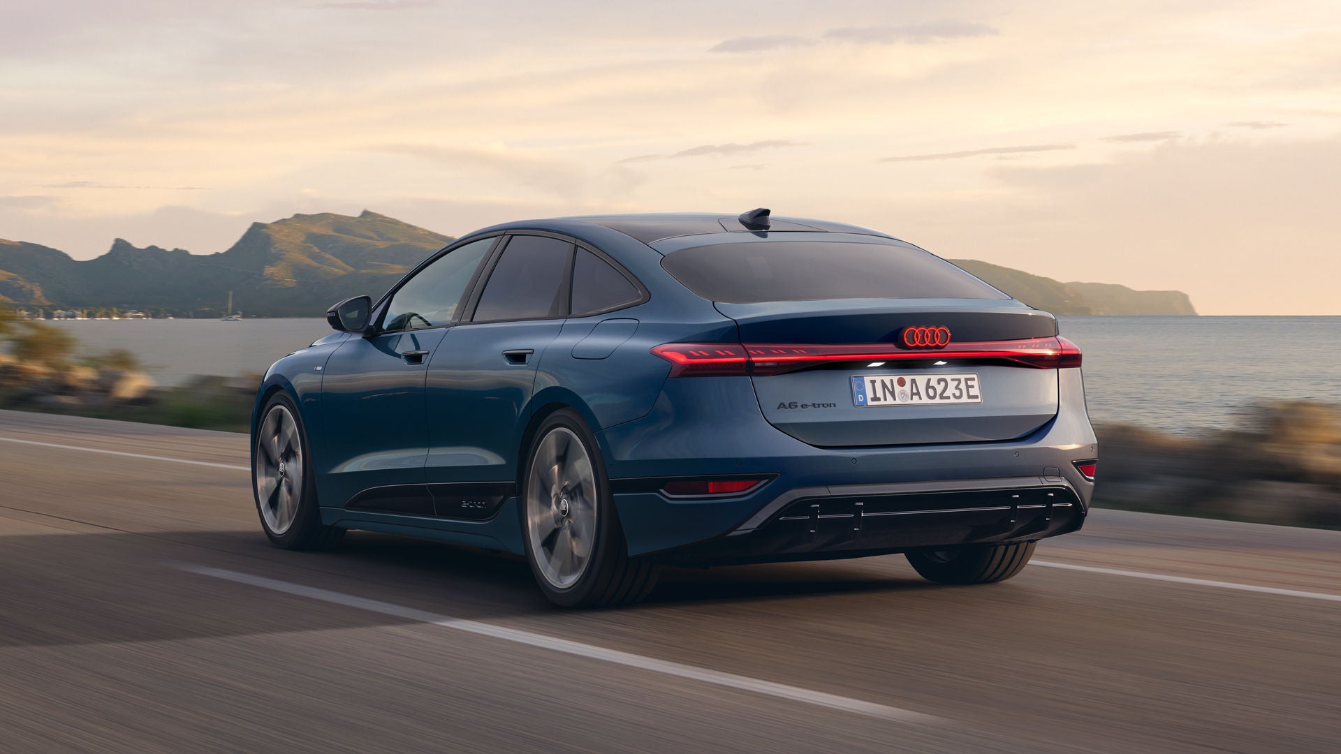 Der A6 Sportback e-tron aus schräger Heckansicht fährt eine Straße parallel zum Ufer entlang. Im Hintergrund sind Berge zu erkennen.