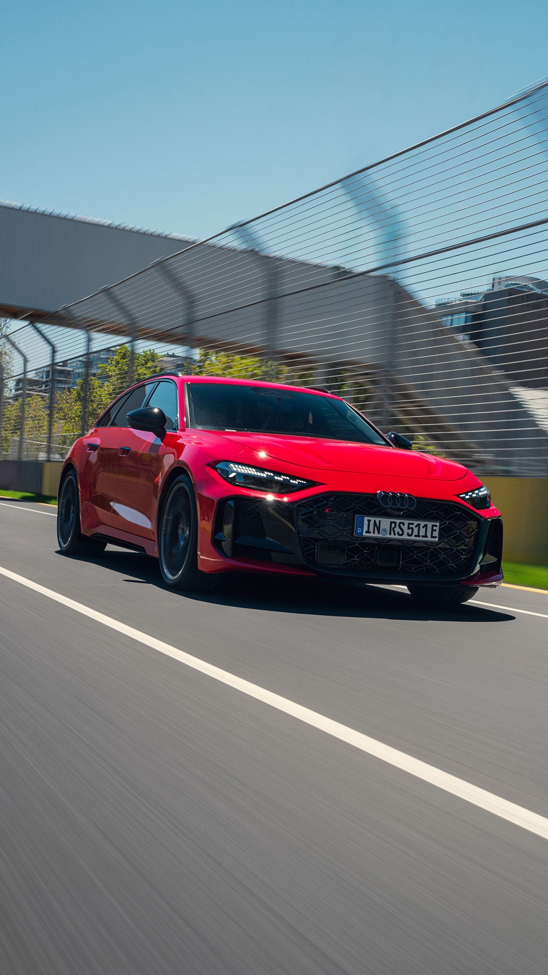 Ein roter Audi RS 5 fährt dynamisch auf der Rennstrecke in Neubug. An der Seite sind Absperrzäune zu erkennen. Im Hintergrund befindet sich in Form einer Brücke ein Übergang über die Rennstrecke, unter der der RS5 gerade durchgefahren ist. 