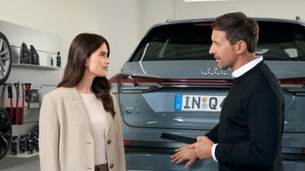 Kundin und Berater sprechen vor dem grauen Audi-Heck im Autohaus; Regale mit Pflegeprodukten im Hintergrund.