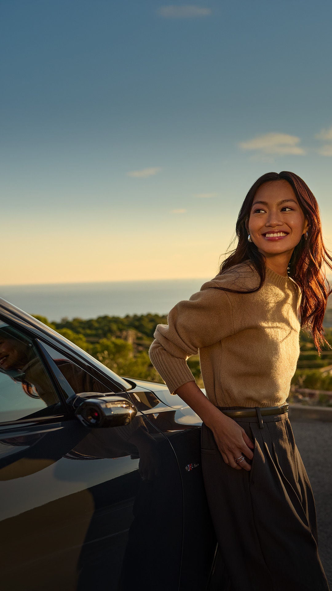 Eine lächelnde Frau lehnt bei Sonnenuntergang an einem dunklen Audi A6 e-tron und blickt über eine Stadt am Meer.