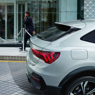 Heckansicht eines silbernen Audi Q3 mit leuchtenden Rücklichtern, geparkt vor einem Gebäude mit Glasfassade und Absperrung.