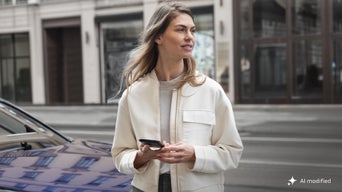 Frau hält ein Smartphone neben einem Auto auf einer städtischen Straße.