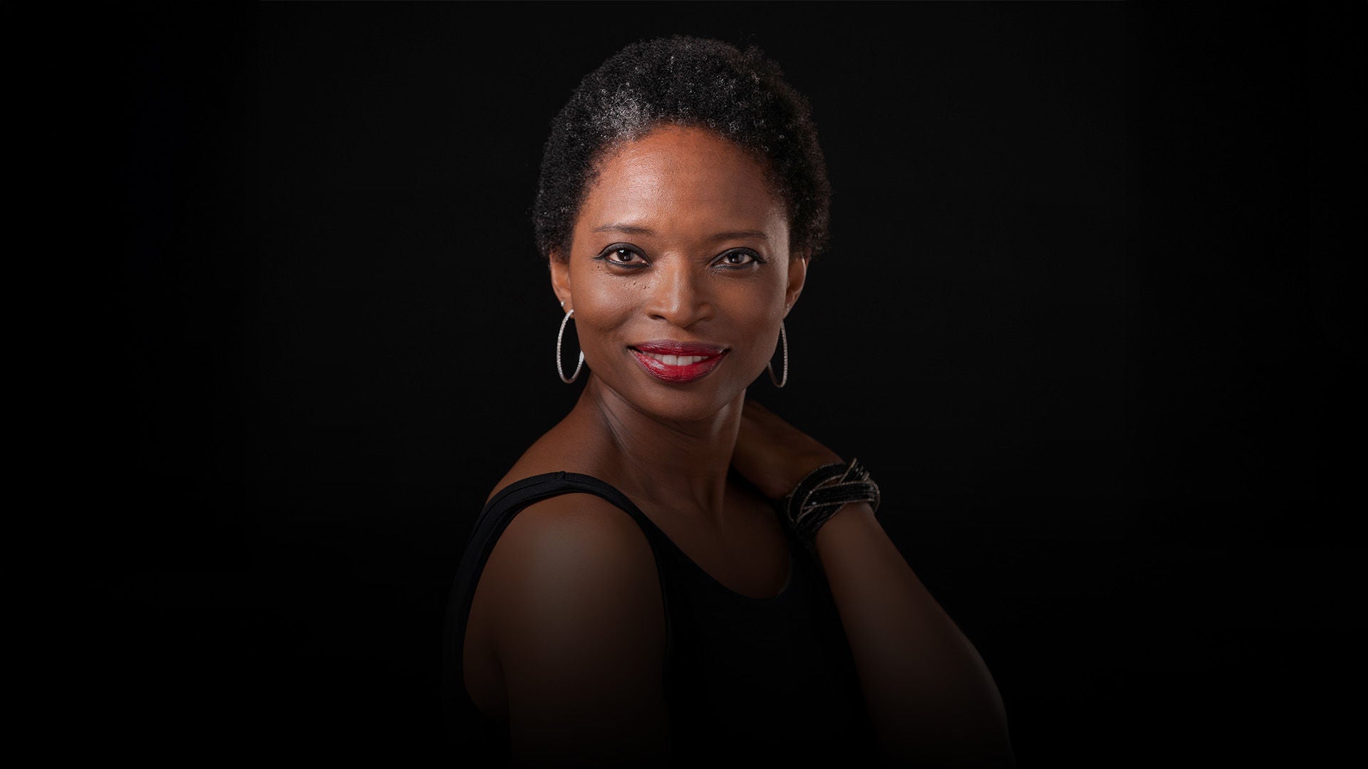 Porträt einer lächelnden Frau vor schwarzem Hintergrund; kurze gelockte Haare, rote Lippen, elegante Ohrringe, ausdrucksstarker Blick.