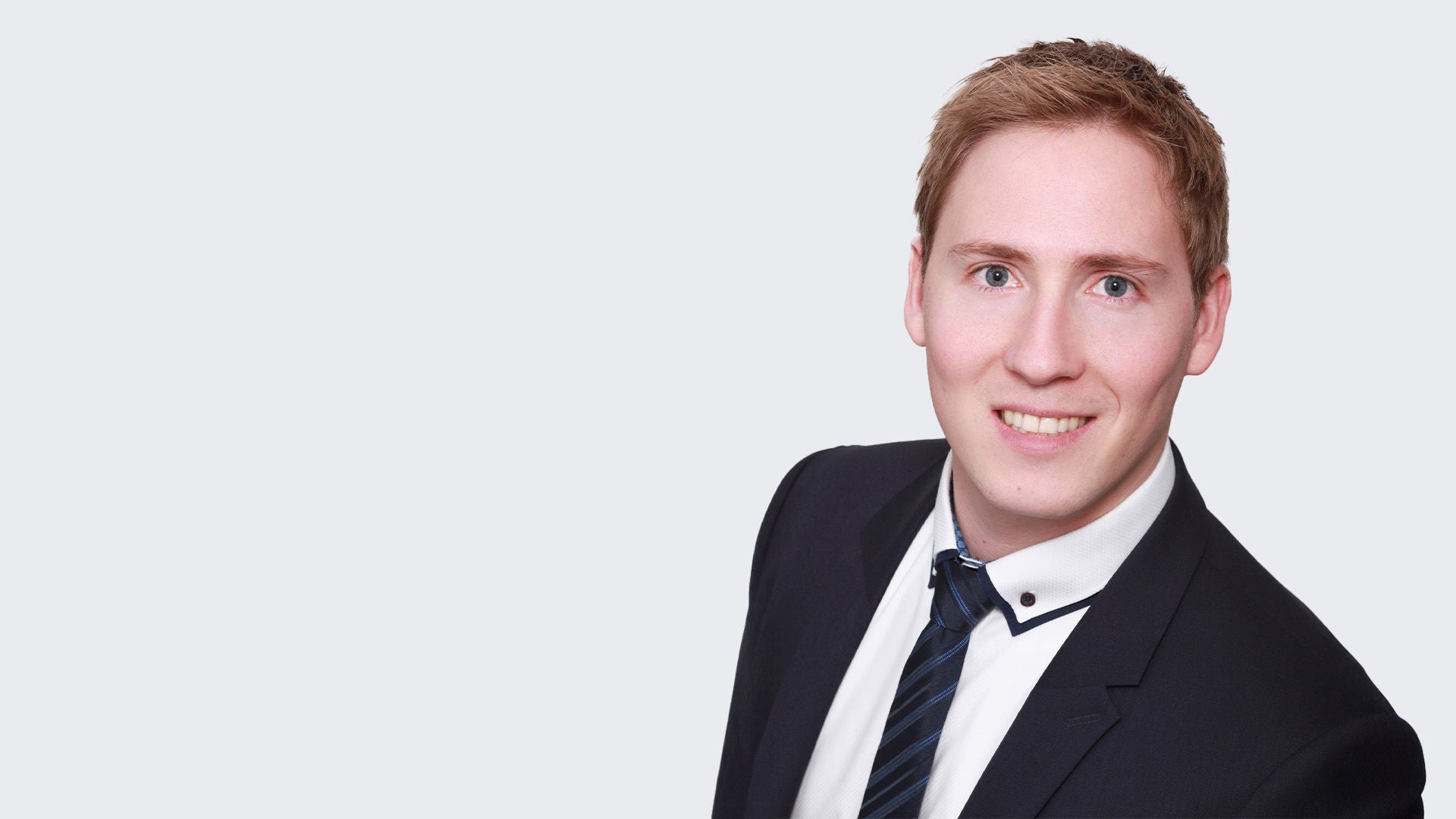 Johannes Roppelt Porträtbild, lächelnd, orange Haare, blaue Augen, schwarzer Anzug, weißes Hemd, dunkle Krawatte, grauer Hintergrund.