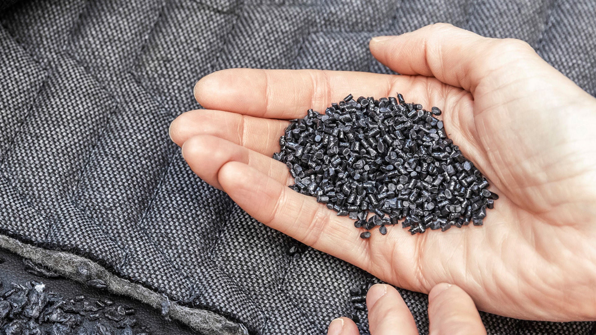 A hand holds dark granules over a dark textile material, surrounded by further granulate.