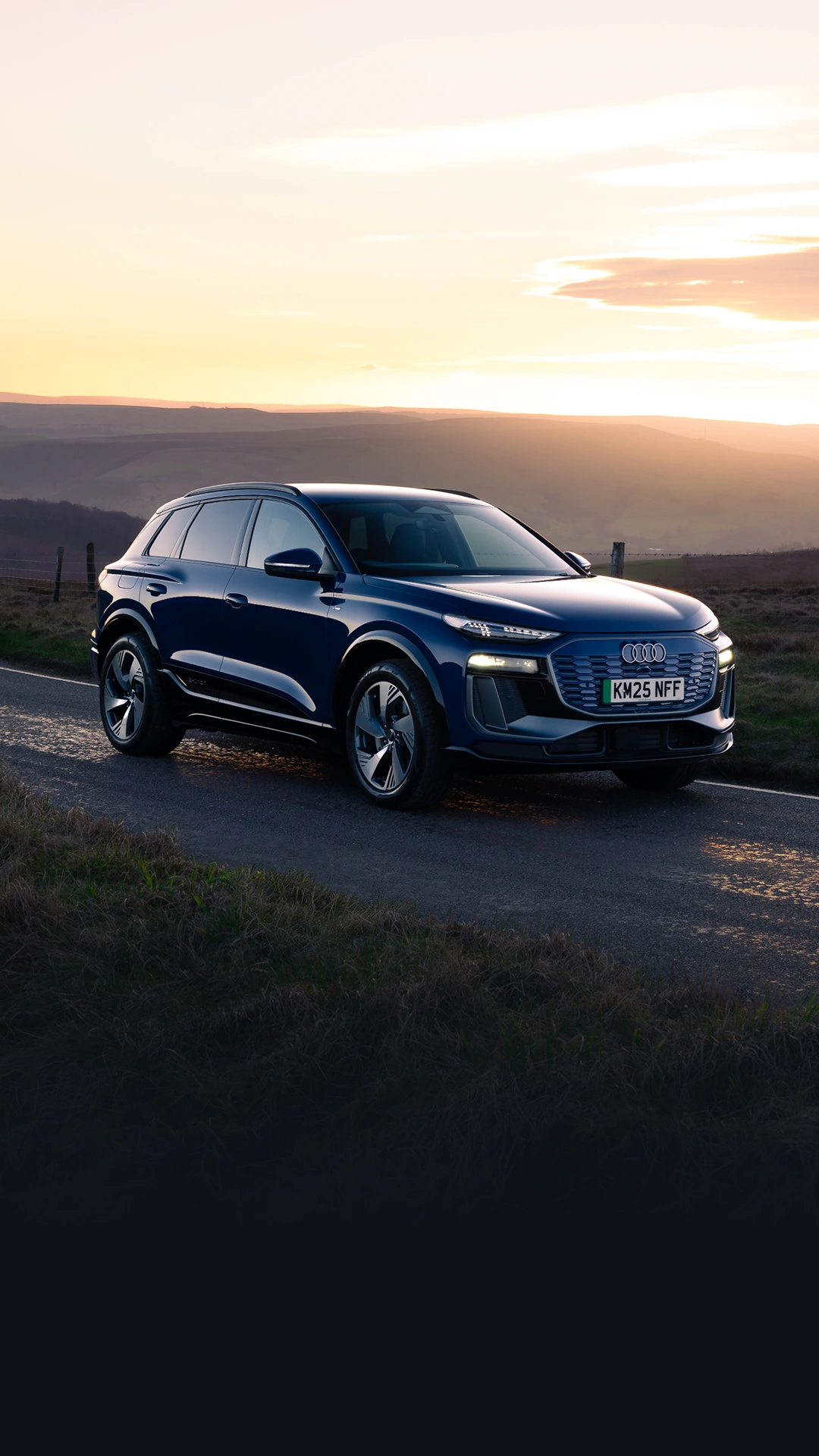Blue Audi Q6 e-tron parked on a rural road at sunset, shown from the front three-quarter angle. The SUV features LED headlights, a wide front grille and aerodynamic alloy wheels. Warm golden light illuminates the surrounding moorland landscape and rolling hills, while reflections shimmer on the road surface beneath the vehicle.