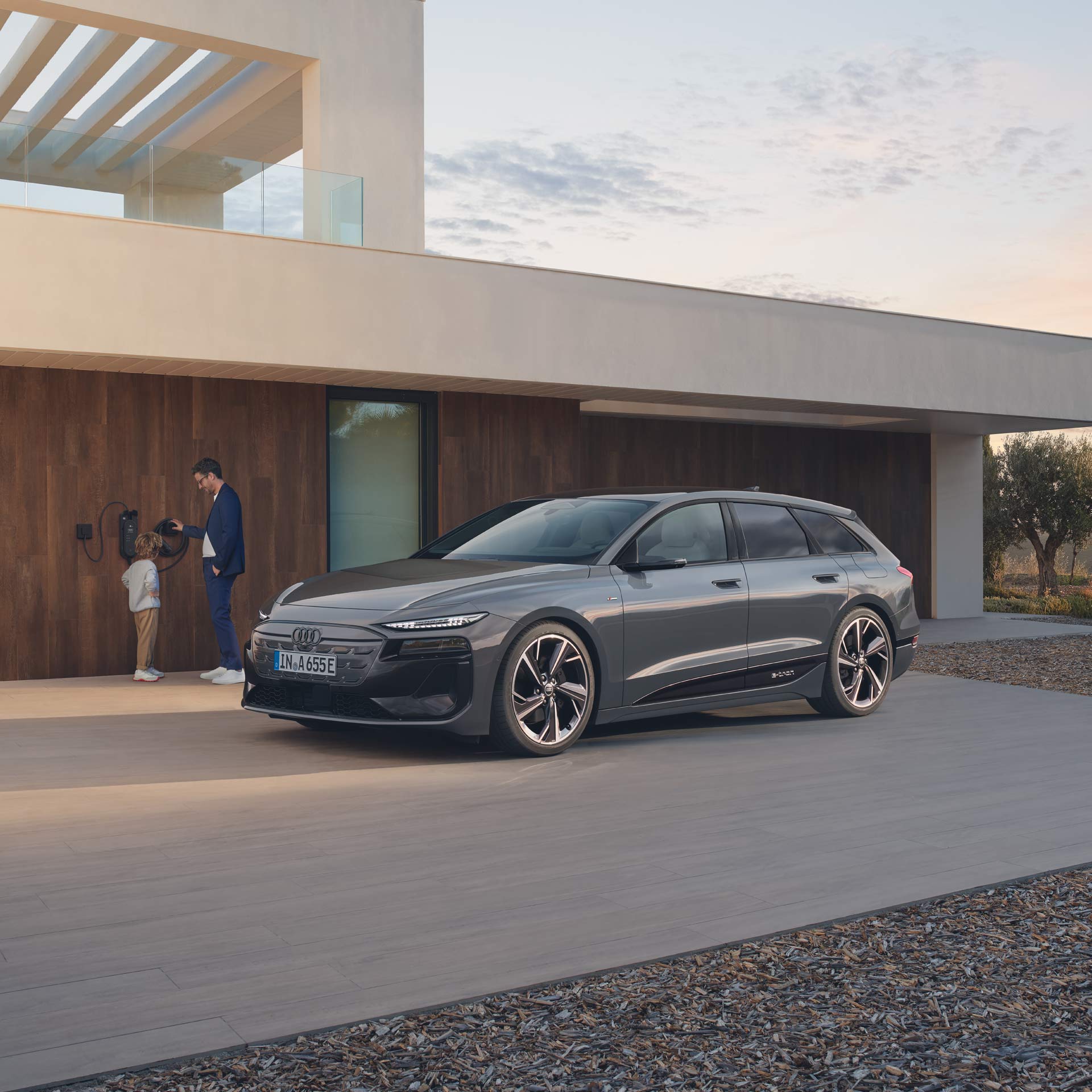 Grauer Audi A6 avant e-tron performance parkt vor Haus, daneben lädt ein Mann mit Kind das Auto an einer Wallbox.
