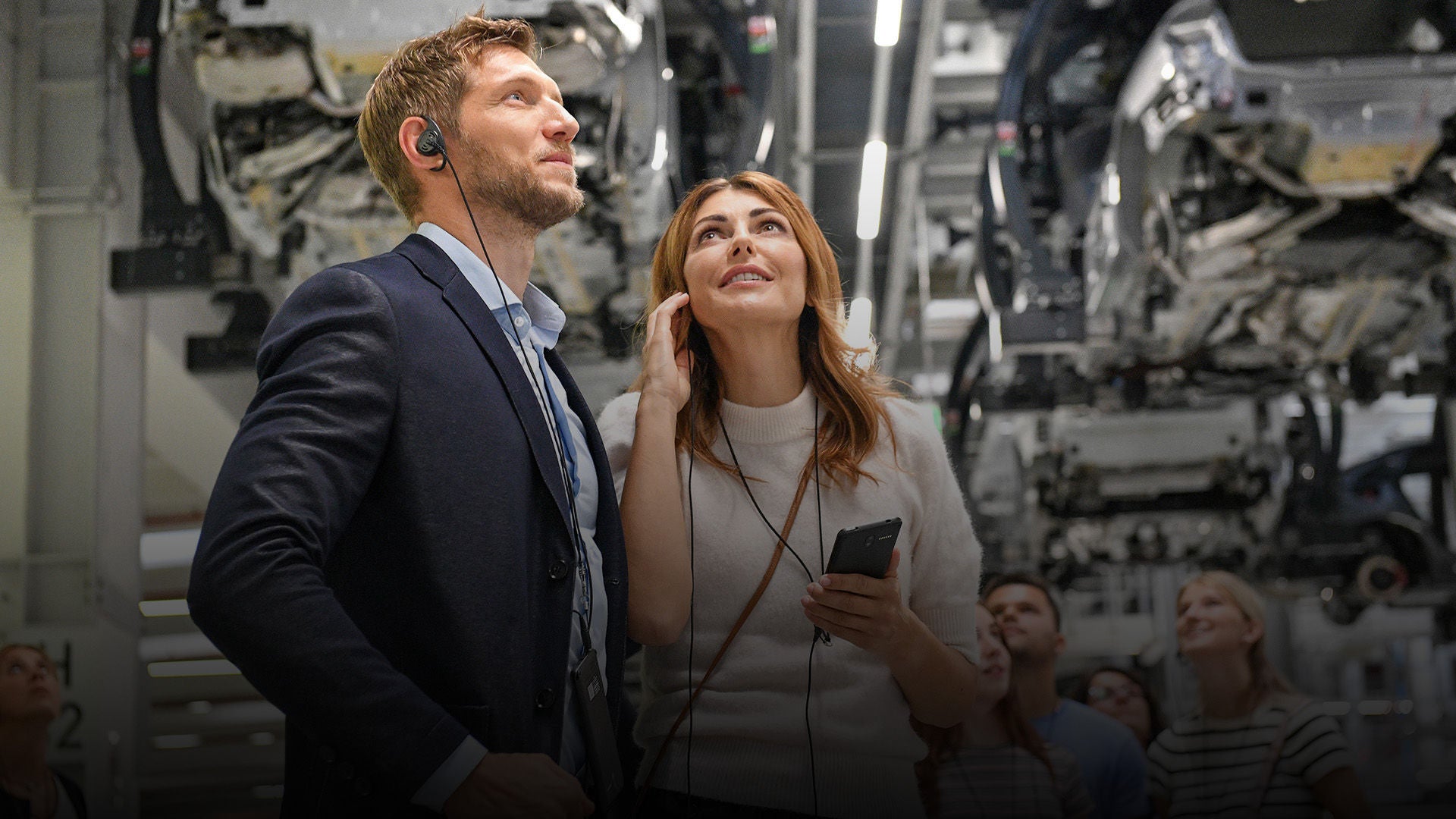 Ein Mann und eine Frau nehmen an einer  Erlebnisführung teil und betrachten die Karosserie eines Audi Modell von unten.