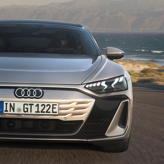 Eine frontale Nahaufnahme des Audi S e-tron GT auf einer Straße an der Küste. Die imposante Lichtsignatur der Scheinwerfer und das sportliche Design stehen im Fokus.