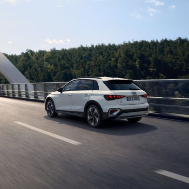 Weißer Audi A3 allstreet TFSI e von schräg hinten auf Brücke, fährt bei Tageslicht vor bewaldetem Hintergrund.