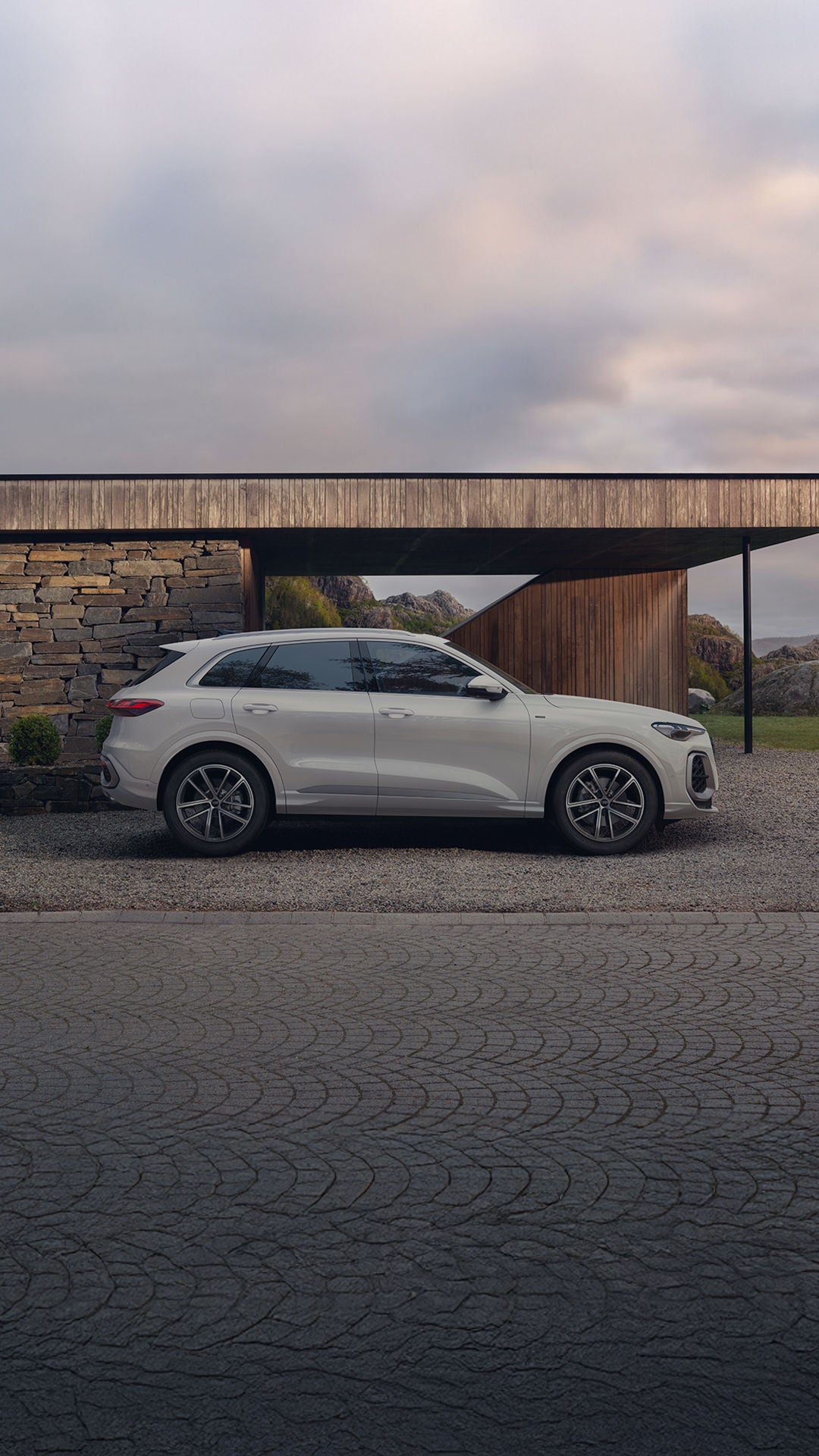 A sleek white SUV parked in front of a modern stone and wood house, surrounded by greenery and rocky hills under a cloudy sky.