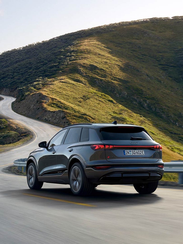 Grauer Audi SQ6 SUV e-tron fährt auf kurviger Landstraße durch hügelige Landschaft, Ansicht vom Fahrzeug hinten links.