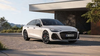 Die A6 Limousine e-hybrid steht auf einem Parkplatz. Im Hintergrund sieht man ein futuristisches Haus.