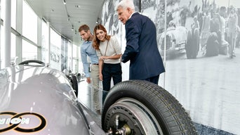 Drei Personen betrachten ein historisches Rennfahrzeug in einer Ausstellung.