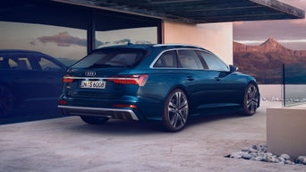 Der S6 Avant schräg von hinten, parkend neben einem modernen Wohngebäude. Die linke Seite des Fahrzeugs spiegelt sich in den bodentiefen Fenstern. Im Hintergrund ein felsiges Gebirge.