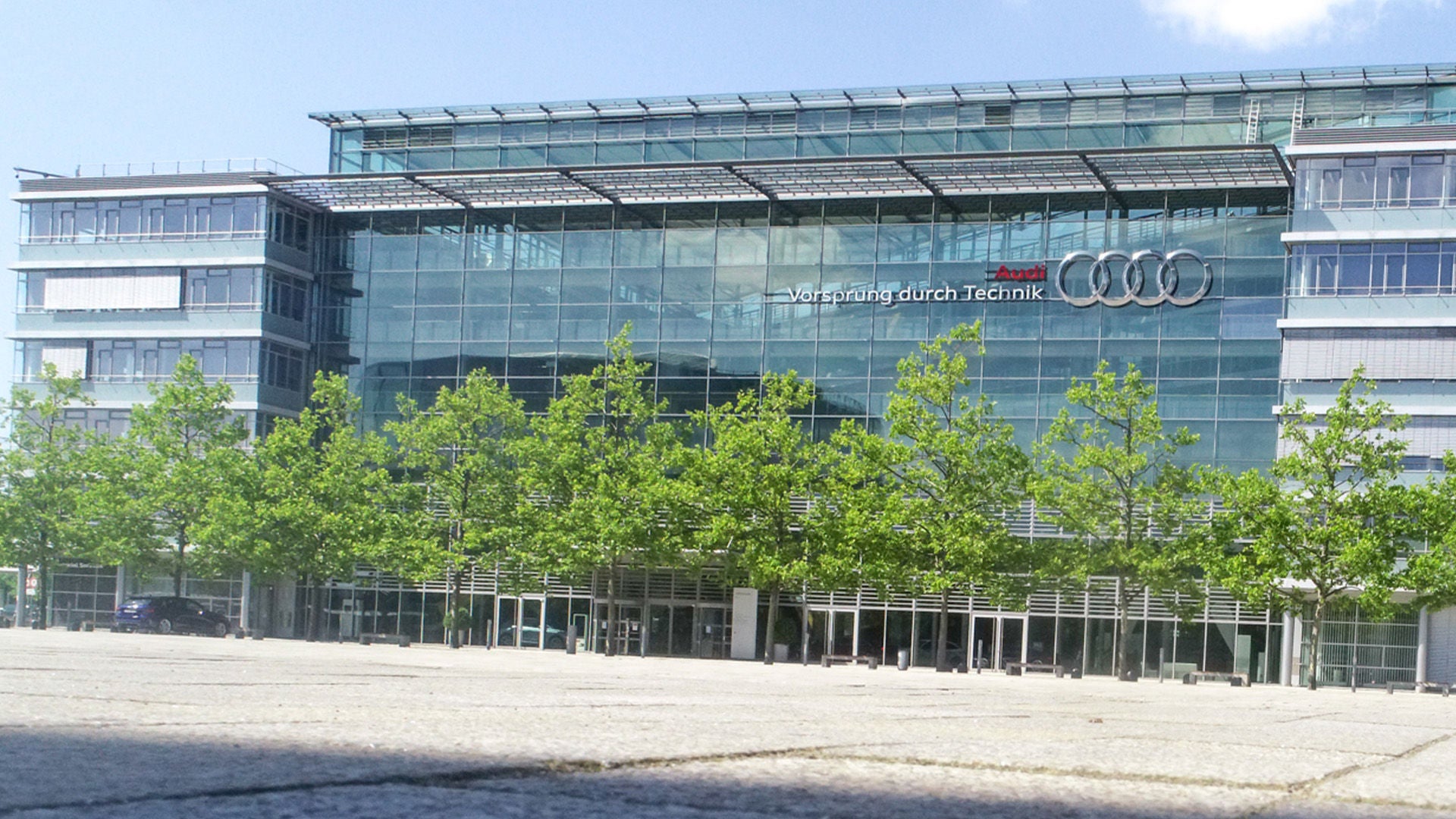 Glass facade of an Audi building with logo, empty space in front of it and row of trees under blue sky.