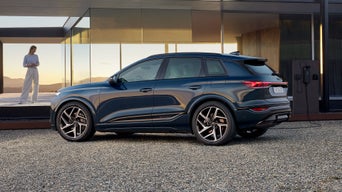 Der Q6 SUV e-tron steht seitlich auf der Kiesfläche vor einem modernen Haus und ist an eine Ladestation angeschlossen. Neben dem Haus steht eine junge Frau.