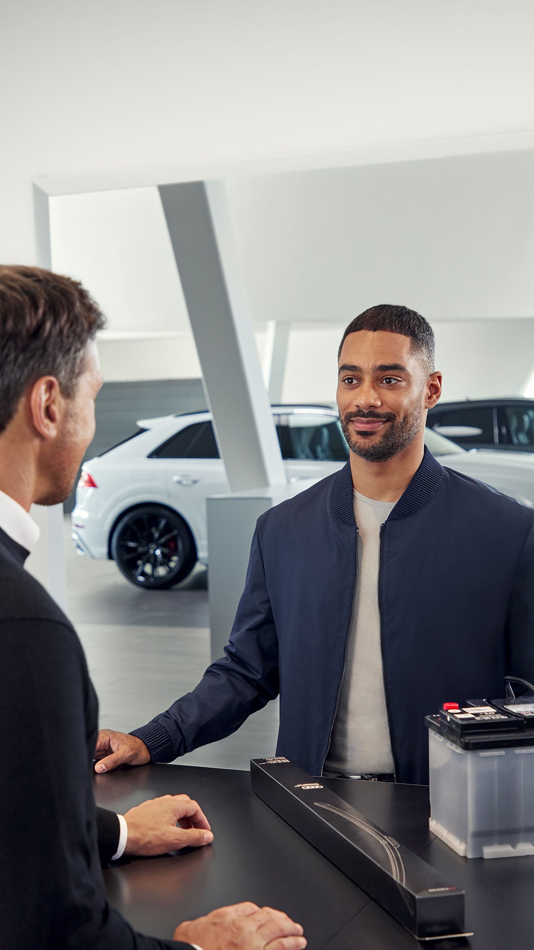 Ein Mann steht an der Kasse eines Autohauses mit einer Autobatterie vor sich. Er wird bedient von einem anderen Mann hinter der Kasse