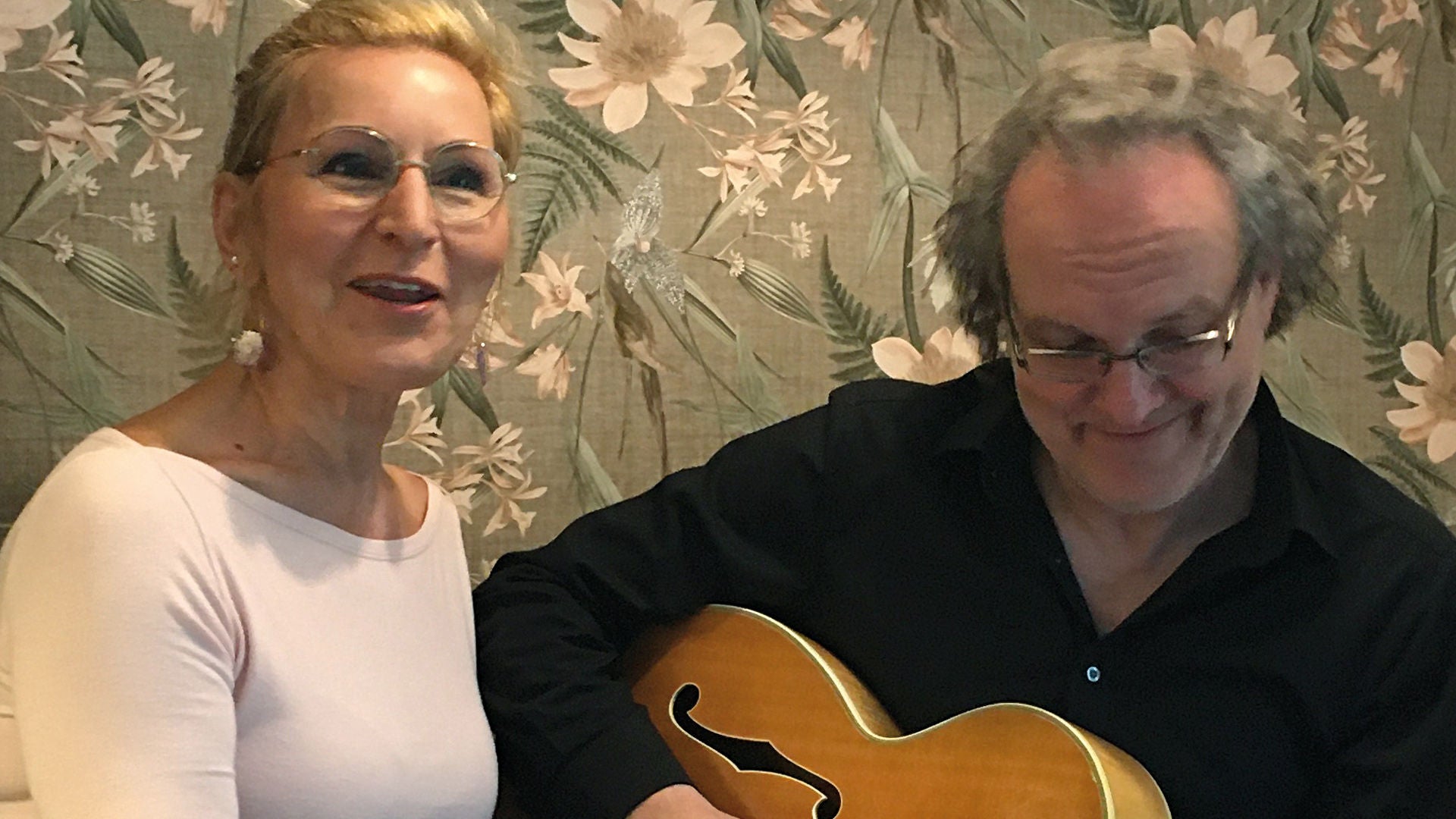 Lächelnde Frau mit blonden Haaren und Brille singt neben Mann mit Gitarre vor geblümter Tapete.