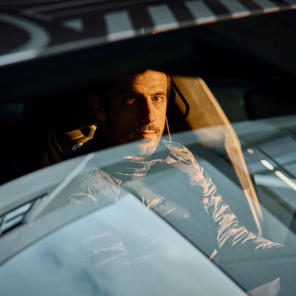 A man stands next to an Audi RS Q8 and gazes into the distance. A skyline can be seen on the horizon.