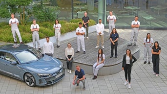 Gruppenfoto von 17 Personen in Berufs- und Freizeitkleidung im Hof neben einem silberblauen Audi vor einem modernen Gebäude.