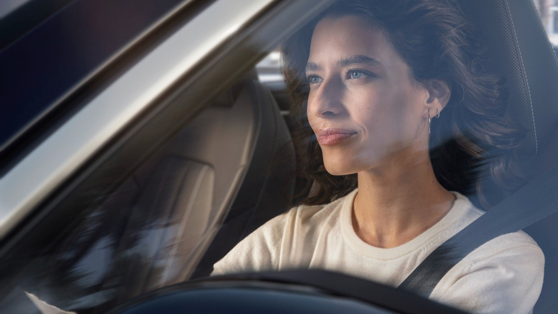A woman with long dark hair sits in a car, gazing thoughtfully out the window. She is wearing a seatbelt.