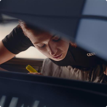 Mechanic, wearing a black t-shirt with Audi logo, inspecting or working underneath a vehicle with a yellow object.