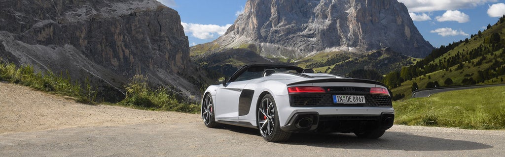 Weißer Audi R8 Spyder V10 performance quattro mit geöffneten Verdeck am Straßenrand vor alpiner Berglandschaft unter blauem Himmel, Rückansicht.