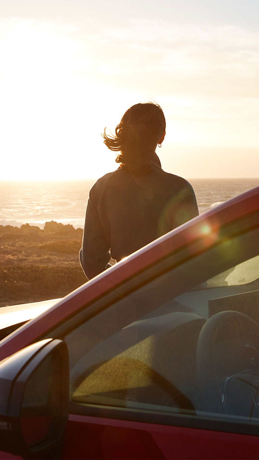 Une femme se tient devant une Audi rouge et regarde le coucher de soleil.