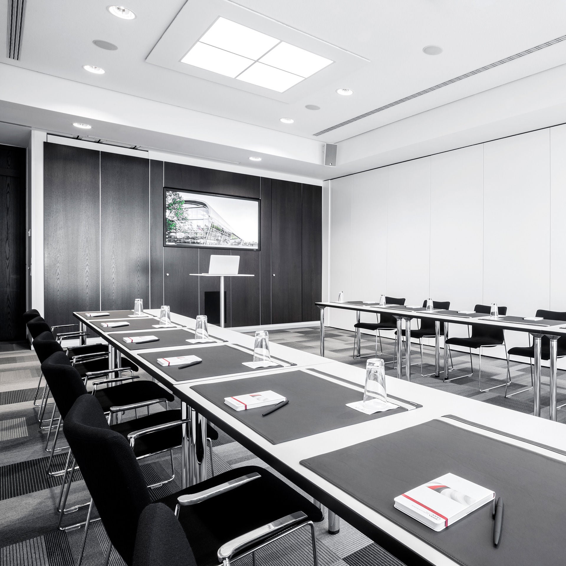 Moderner Konferenzraum mit langen Tischen, bestuhlten Sitzreihen, Schreibblöcken, Wassergläsern und Präsentationsbildschirm.