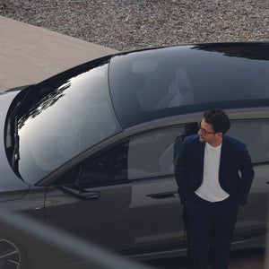 A man in a suit stands beside a sleek, dark car with a reflective sunroof. He looks thoughtfully to the side. The scene conveys elegance and contemplation.
