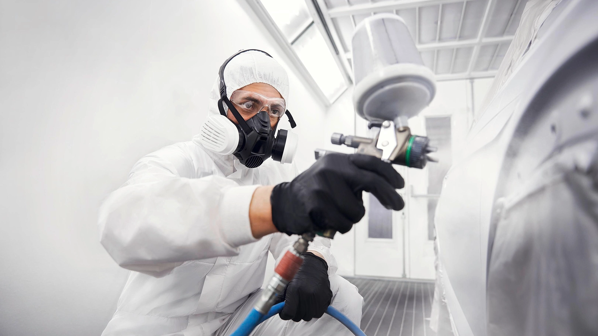 Technician wearing protective equipment applying paint to a vehicle panel in a professional spray booth.