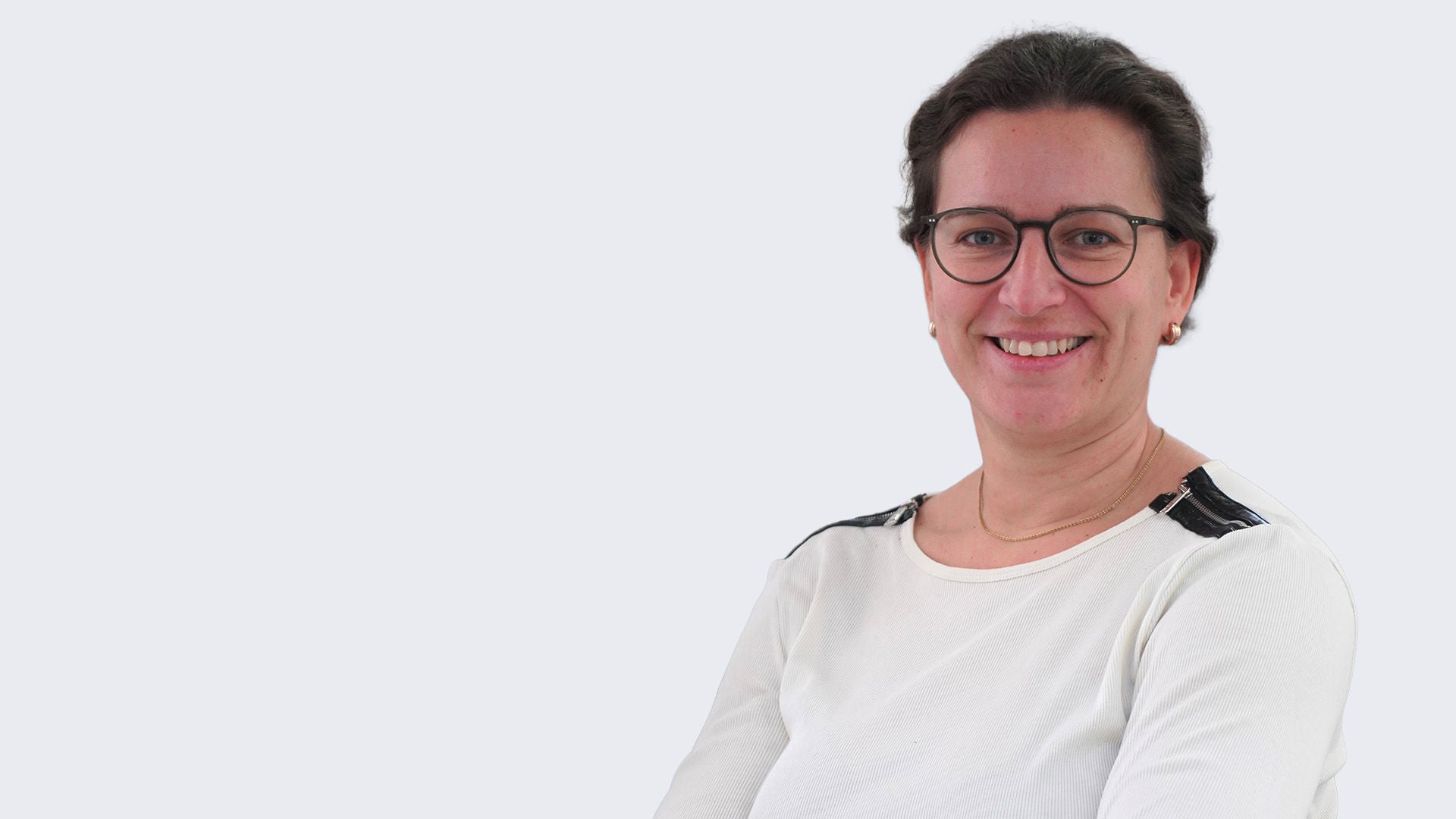 Porträt einer lächelnden Frau mit Brille und weißem Oberteil vor hellem Hintergrund.
