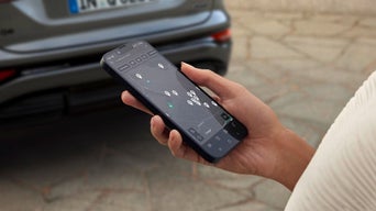 Hand hält Smartphone mit Ladepunktsuche, im Hintergrund das Heck eines Audi-Fahrzeugs.