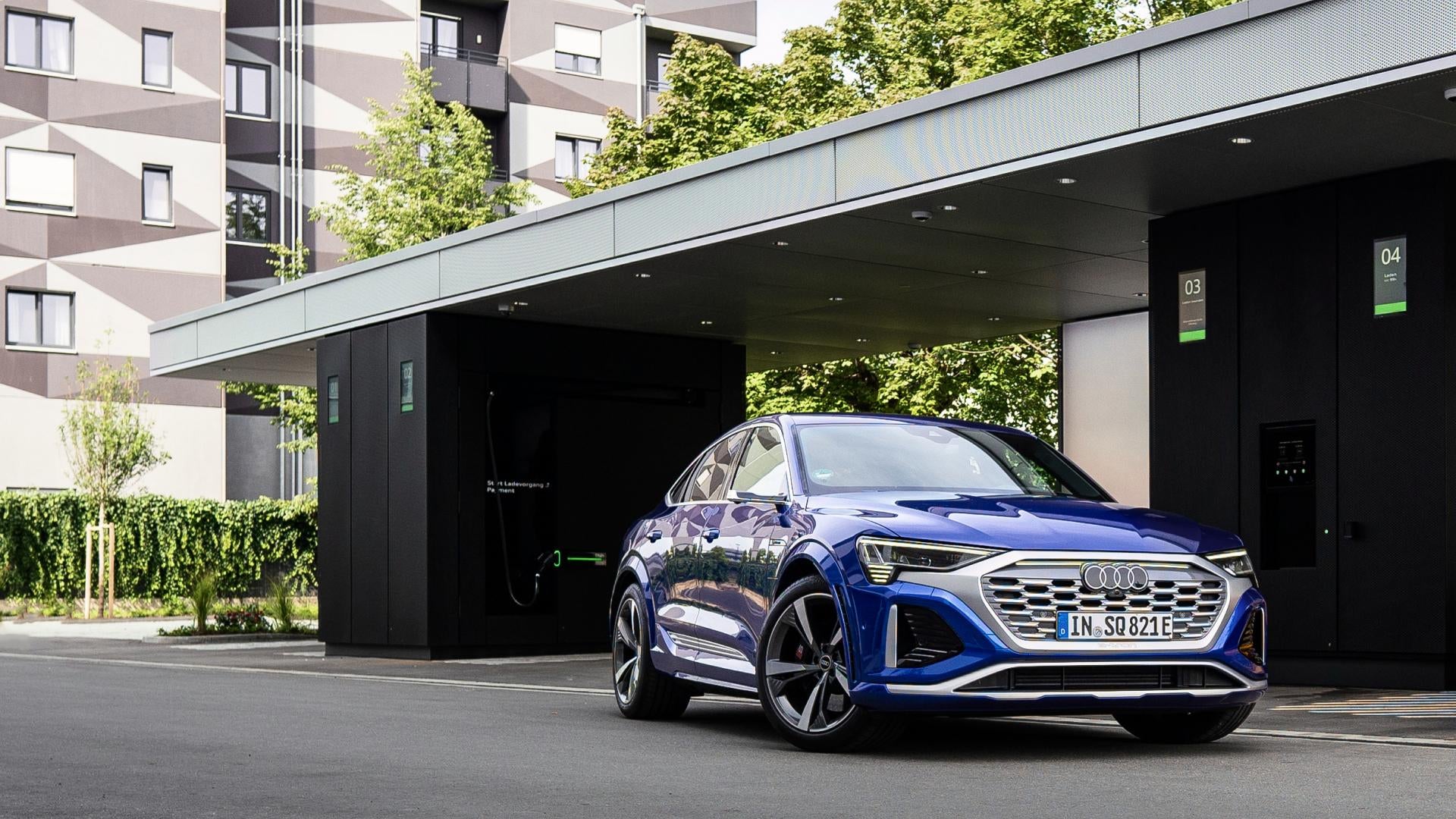 Blauer Audi e-tron parkt vor einer modernen Audi-Ladestation mit zwei Ladepunkten in städtischer Umgebung.