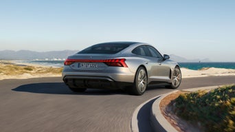 Ein silberner Audi S e-tron GT fährt auf einer kurvigen Küstenstraße mit Blick auf das Meer und eine Stadt im Hintergrund.