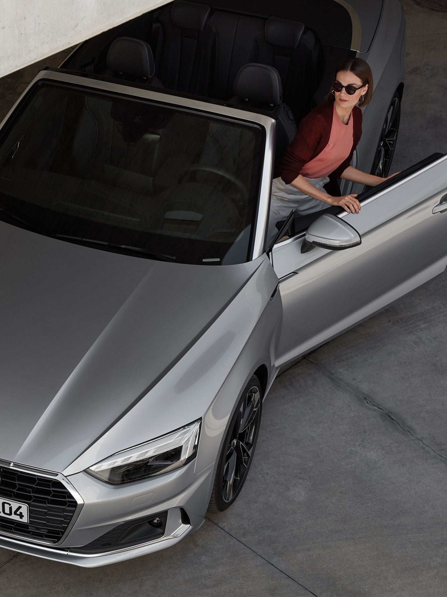 A person in a red dress stands beside a silver Audi car near a futuristic building with a unique texture.