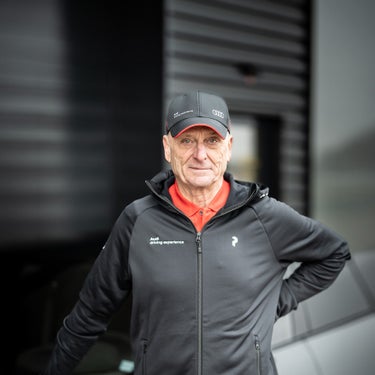 Porträt eines Mannes mit schwarzer Kappe und Audi-Jacke vor dunklem Gebäudehintergrund.