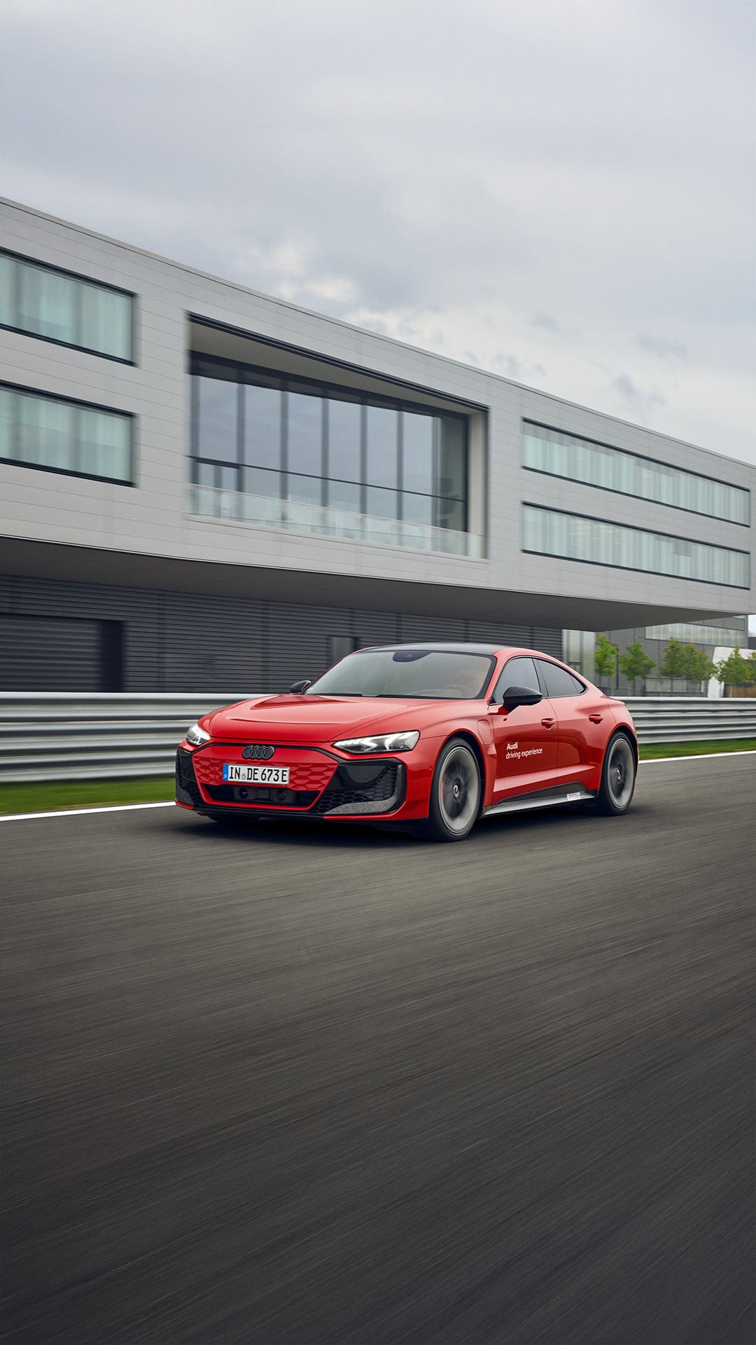 Roter Audi e-tron GT fährt auf Rennstrecke vor modernem Gebäude mit Glasfassade entlang.
