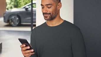 Um homem olha para a tela do celular. Ao fundo, há um Audi estacionado.