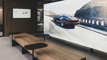 Moderner Sitzbereich mit langen Holzbänken und integrierten Tischen, im Hintergrund großer Bildschirm mit Audi-Fahrzeug.