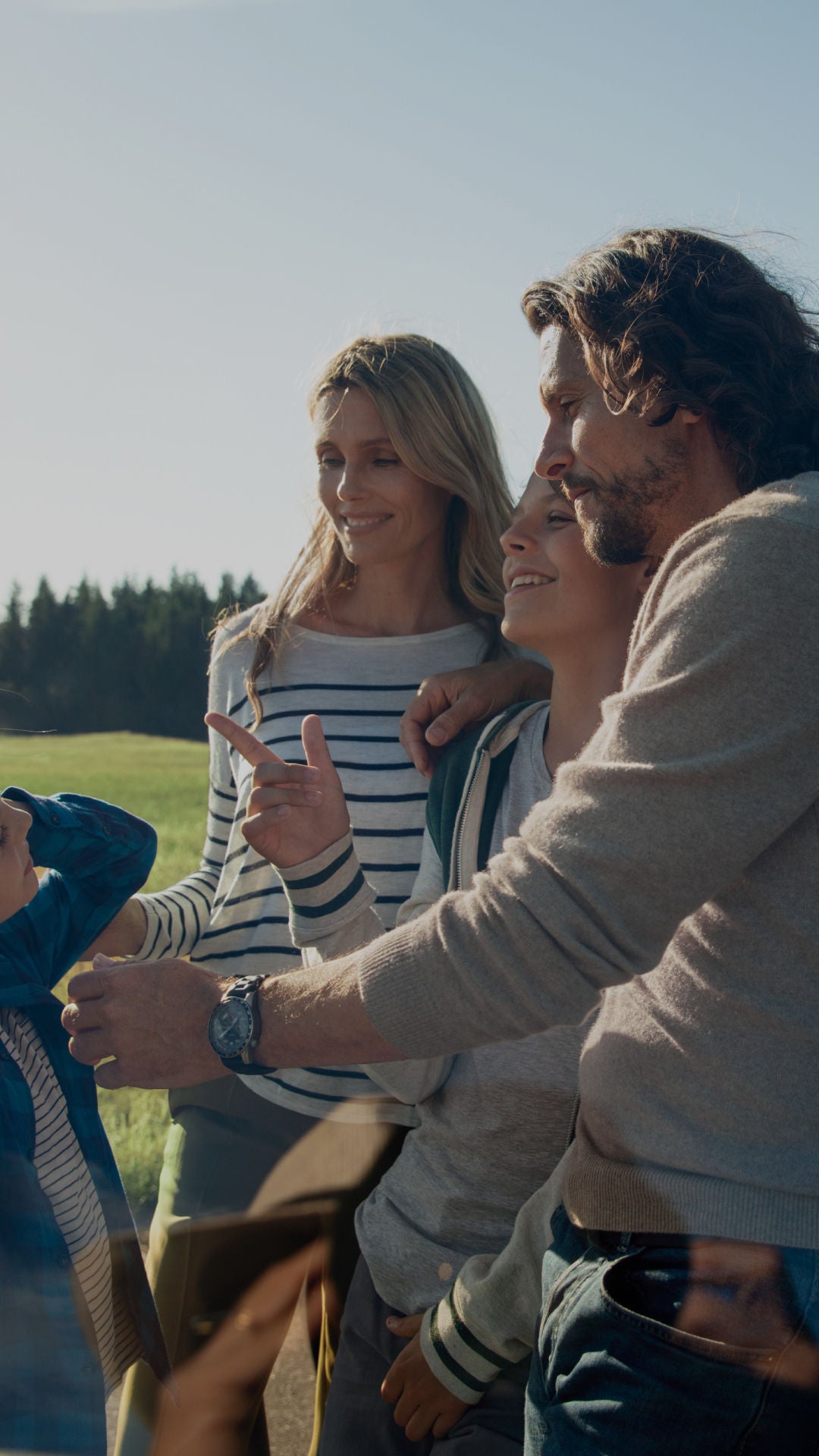 A group of people stands outside a car in a bright, sunny field, engaged in conversation, with trees in the background.