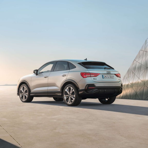 Rear angled side view of silver Audi Q3 Sportback with metal structure building in the background.