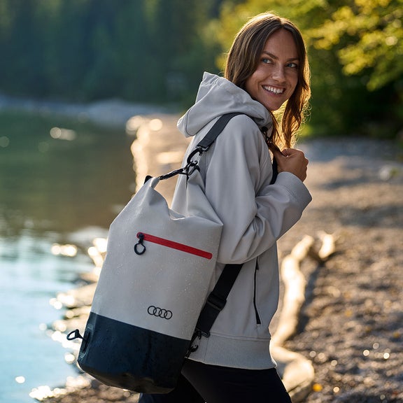 A woman walks along a lake. She looks into the camera. She is carrying a duffel bag with the Audi emblem over her shoulder.