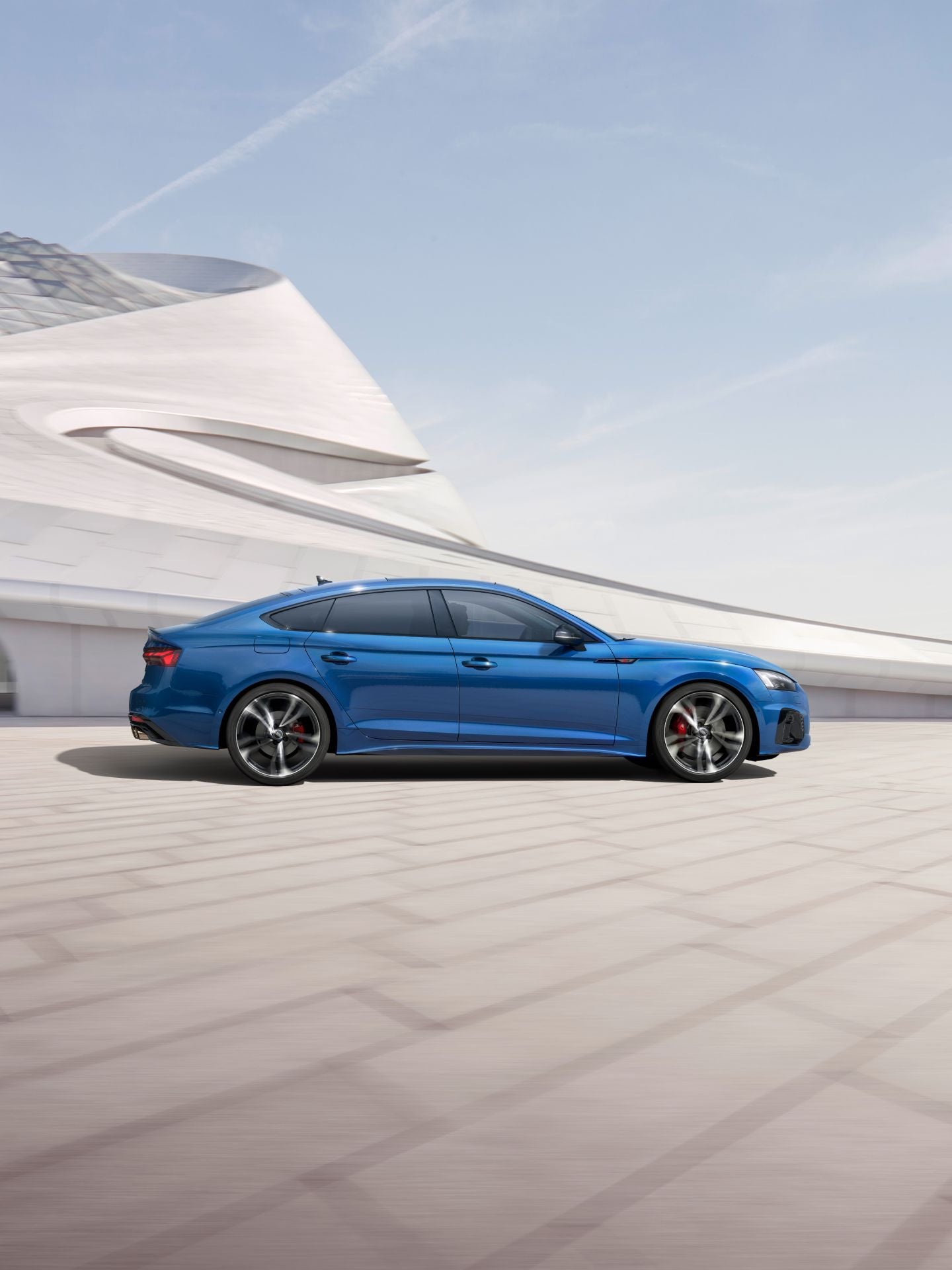 A blue Audi S5 Sportback driving past a modern white building with a sleek design.