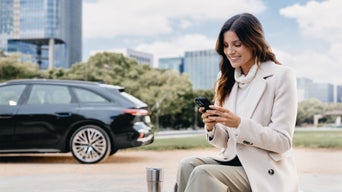 Une femme souriante est assise à l'extérieur, vêtue d'un manteau blanc et utilisant un smartphone. À l'arrière-plan, une Audi noire est garée devant un décor urbain avec des arbres et des immeubles de bureaux.