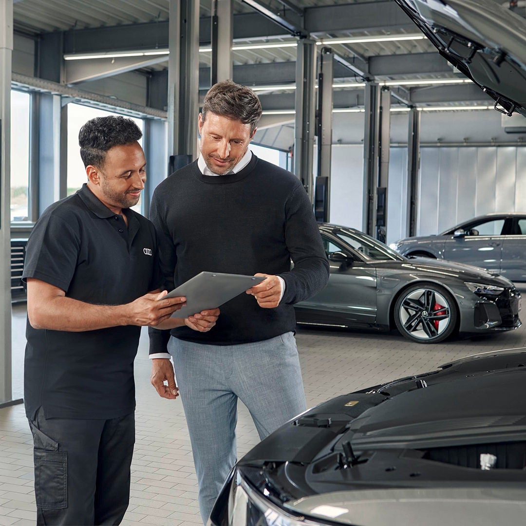 Servicemitarbeiter zeigt Kunden ein Tablet vor geöffnetem Motorraum in heller Werkstatt mit zwei geparkten Audi-Fahrzeugen.